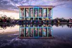 National Library of Australia, eastern facade, with illuminated projections of their Australian history photographic collection, at dusk, with exact mirror image reflection in the forecourt pond. People are sitting on the edge of the pond admiring the view. Vertical columns are picked out in light blue. - Enlighten